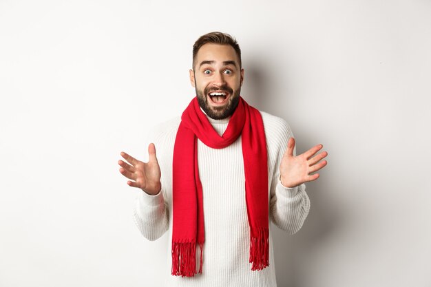 Weihnachtsferien und Feierkonzept. Bärtiger Kerl, der überrascht auf Neujahrs-Promo-Angebote schaut, erstaunt nach Luft schnappt, roten Schal und Pullover trägt, weißer Hintergrund.