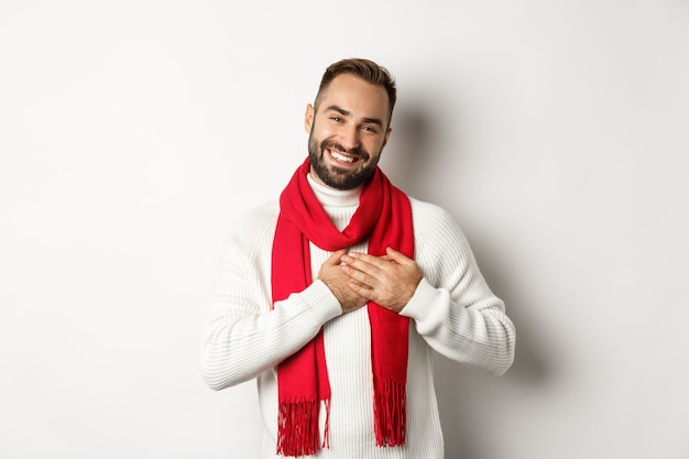 Weihnachtsferien und Feierkonzept. Gut aussehender Mann, der sich für Geschenke des neuen Jahres bedankt, die Hände am Herzen hält und dankbar lächelt, vor weißem Hintergrund stehend.