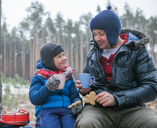 Foto weihnachtsferien, vater und sohn trinken heißes neujahrsgetränk. glückliche familie bei einem spaziergang im freien im sonnigen winterwald