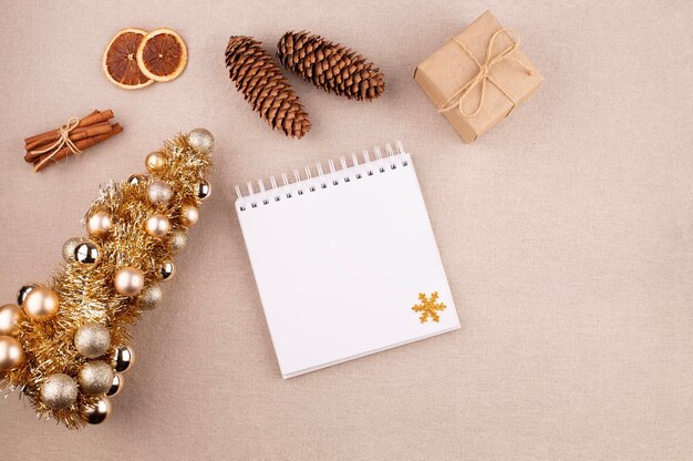 Weihnachtsferienhintergrund mit Tagebuch und goldfarbenem Baum Zimtstäbchen und Geschenk in Kraftpapier gewickelt getrocknete Orangen auf Leinstoffhintergrund flacher Kopierraum