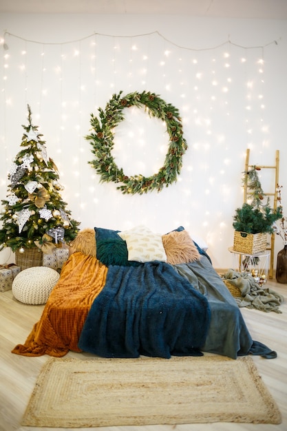 Weihnachtsfestliches Interieur in grauen und weißen Farben. An der Wand hängt ein runder grüner Kranz. Ein mit kuscheligen Decken bedecktes Bett steht in einem mit einer Girlande geschmückten Haus.