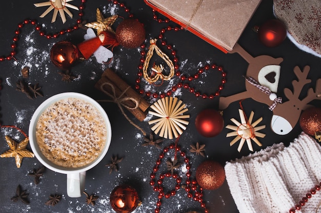 Weihnachtsflache mit heißem Kaffee oder Kakao auf schwarzem Holzhintergrund mit handgemachtem Weihnachtsspielzeug, roten Kugeln, Perlen, Nelken und Zimt. Nahaufnahme, Ansicht von oben