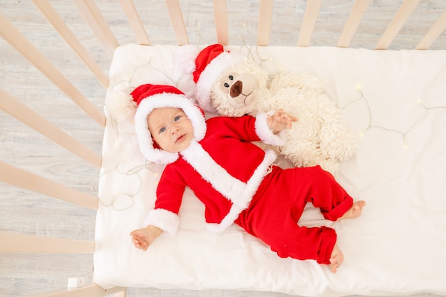 Weihnachtsfoto eines Babys in einem Weihnachtsmannkostüm, das in einem Kinderbett zu Hause mit einem Spielzeug in einer Weihnachtsmannmütze liegt