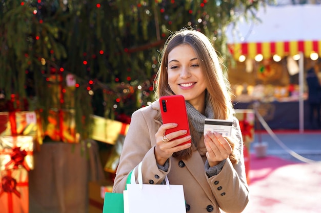 Weihnachtsfrau, die Einkaufstaschen im Freien trägt Schönes lächelndes Mädchen, das ihre Geschenke online mit Handy und Kreditkarte bezahlt, mit Weihnachtsmärkten im Hintergrund