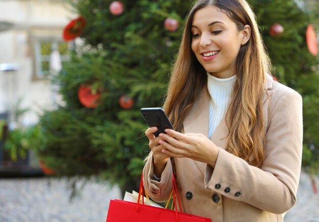 Weihnachtsfrau, die online auf dem Smartphone in der Straße mit Weihnachtsbaum auf Hintergrund kauft.