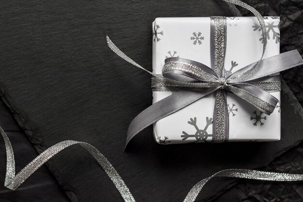 Weihnachtsgeschenkbox, eingewickelt in ein weißes Papier mit Schneeflocken und mit einem Band auf dunklem Hintergrund gebunden. Flache Lage, Ansicht von oben, Kopienraum