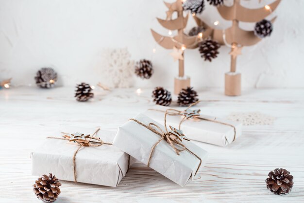 Weihnachtsgeschenke verpackt in Bastelpapier mit Bindfäden auf dem Hintergrund von Weihnachtstannen aus Pappe auf einem Holztisch. Organisation eines umweltfreundlichen Urlaubs. Platz kopieren