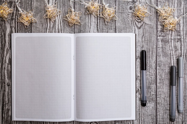 Weihnachtsgirlande, leeres quadratisches Notizbuch, Stifte auf einem grauen Holztisch
