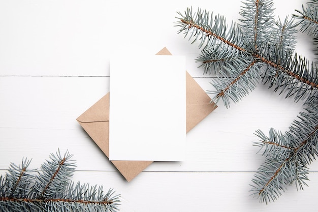 Weihnachtsgrußkarte Mockup mit Kraftpapierumschlag und frischem grünen Tannenzweig auf weißem Holzhintergrund Draufsicht flach lag Leere Winterurlaubskarte