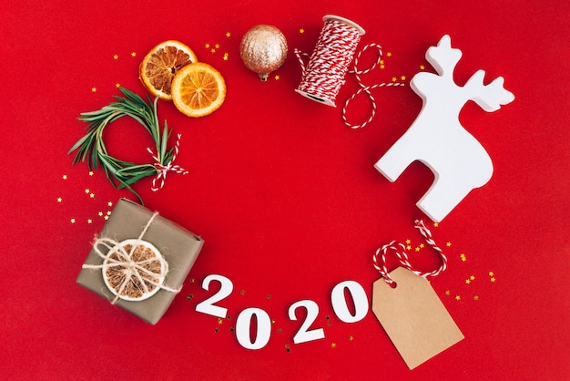 Weihnachtshandgemachter ovaler Rahmen gemacht von 2020 hölzernen Ziffern und von Tannenbaumasten und von anderen Weihnachtsdekorationen auf rotem Hintergrund.