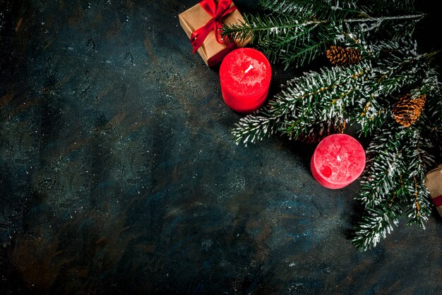 Weihnachtshintergrund für Grußkarte. Weihnachtsbaumaste mit Schneeeffekt mit festlichem rotem Band, Kiefernkegeln, Geschenkboxen und Kerzen, auf Draufsicht des dunkelblauen Hintergrundes