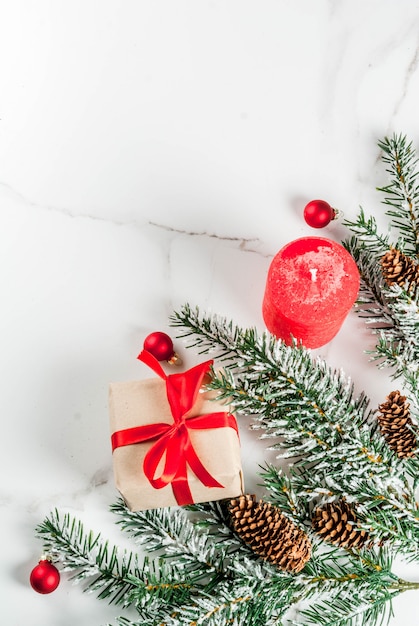 Weihnachtshintergrund für Grußkarte. Weihnachtsbaumaste mit Schneeeffekt mit festlichem rotem Band, Kiefernkegeln, Geschenkboxen und Kerzen, auf weißem Marmorhintergrund