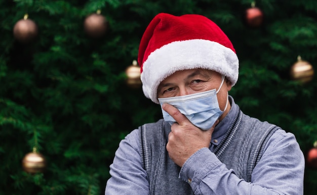 Weihnachtsidee. Schließen Sie herauf Porträt des älteren Mannes, der einen Weihnachtsmannhut und eine medizinische Maske mit Emotion trägt. Vor dem Hintergrund eines Weihnachtsbaumes. Coronavirus Pandemie