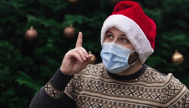 Weihnachtsidee. Schließen Sie herauf Porträt des Mannes, der einen Weihnachtsmannhut, Weihnachtspullover und medizinische Maske mit Emotion trägt. Vor dem Hintergrund eines Weihnachtsbaumes. Coronavirus Pandemie