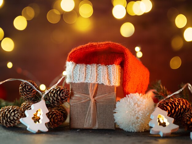 Weihnachtskarte. Eine Geschenkbox in einer gestrickten Weihnachtsmannmütze auf einem Hintergrund mit einer leuchtenden Bokeh-Girlande.