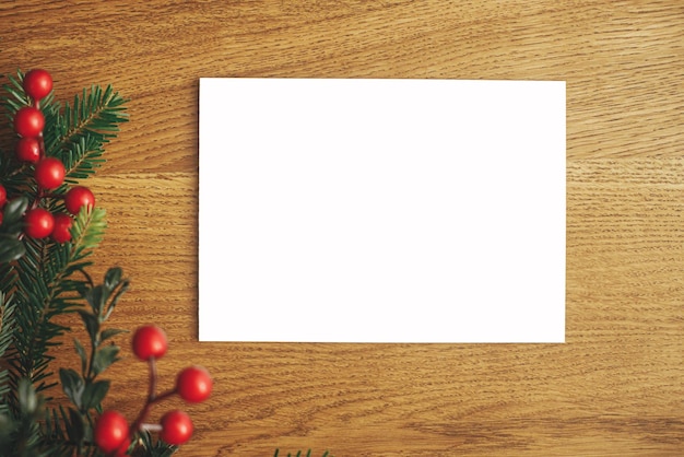 Weihnachtskarten-Attrappe flach liegend Leere Grußkarte und Tannenzweige mit roten Beeren auf Holztisch Platz für Text Postkartenvorlage für Weihnachtsgrüße