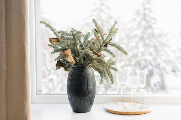 Weihnachtskomposition aus Weihnachtsbaumzweigen in einer schwarzen Vase am Fenster eines gemütlichen Hauses