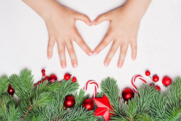 Weihnachtskomposition mit Kinderhänden, die Herz grün machen Xmas Tannenzweig rote Beeren der Stechpalme und Kugeln auf weißem Hintergrund Buntes Weihnachtskonzept Draufsicht flach lag mit Kopierraum