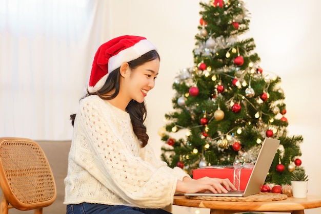 Weihnachtskonzept Asiatische Frau, die neben dem Weihnachtsbaum sitzt und online auf dem Laptop arbeitet und einkauft