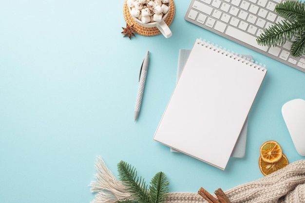 Weihnachtskonzept Draufsichtfoto von Notizblock, Stift, Tastatur, Tasse Kakao mit Marshmallow-Strickschal, Tannenzweigen, getrockneten Zitrusscheiben und Zimt auf isoliertem pastellblauem Hintergrund mit Leerraum