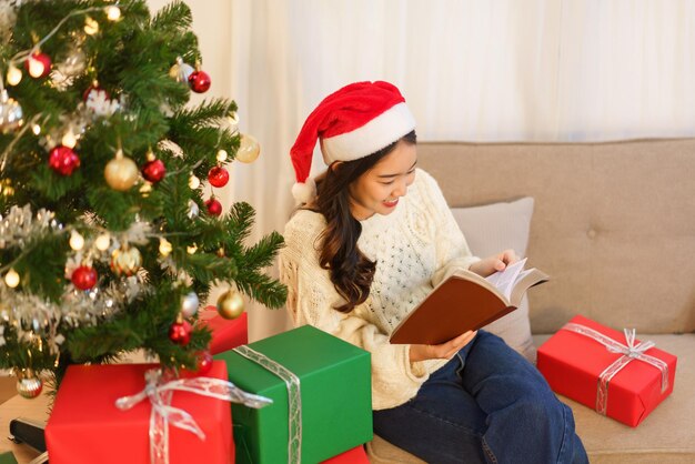 Weihnachtskonzept Junge asiatische Frau, die auf der Couch sitzt und ein Buch in der Nähe des Weihnachtsbaums liest