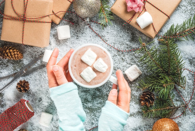 Weihnachtskonzept, Tabellenhintergrund mit Schnee, Weihnachtsbaumaste, Mädchen hält Becheresprit-Eibisch der heißen Schokolade, Geschenke oder Geschenke, Kiefernkegel und Dekoration