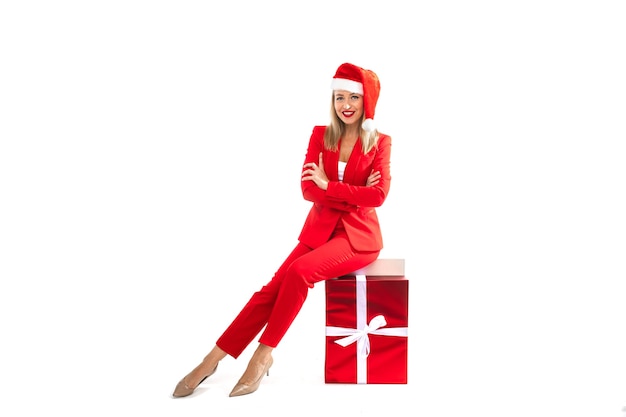 Weihnachtskonzeptfoto der eleganten blonden Dame im roten Anzug und in den hohen Absätzen, die auf verpackter Geschenkbox sitzen. Winterferienkonzept