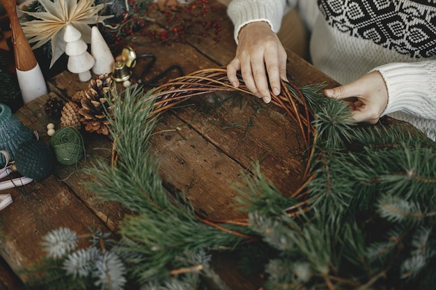 Weihnachtskranz machen Frau, die Tannenzweige hält und Weihnachtskranz auf rustikalem Hintergrund arrangiert