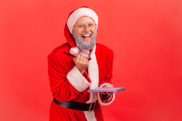 Weihnachtsmann hält Papiernotizbuch und Stift in den Händen, lacht und schaut in die Kamera.