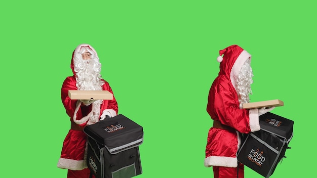 Weihnachtsmann liefert Pizzakarton aus Thermorucksack, Person porträtiert festlichen traditionellen Charakter in Kostüm vor Greenscreen-Hintergrund. Winterfeier, Lieferbote.