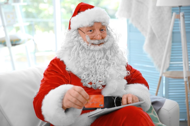 Weihnachtsmann mit Kreditkarte und Tablet auf der Couch zu Hause