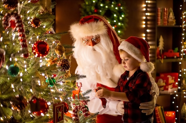 Weihnachtsmann schmückt Weihnachtsbaum mit kleinem Jungenassistenten zu Hause Neujahr glückliche Kindheit