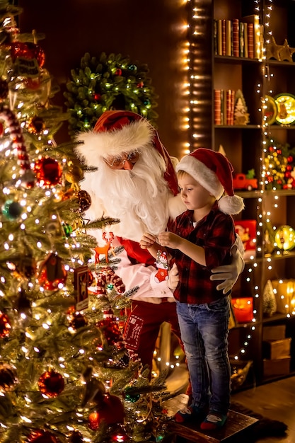 Weihnachtsmann schmückt Weihnachtsbaum mit kleinem Jungenassistenten zu Hause Neujahr glückliche Kindheit