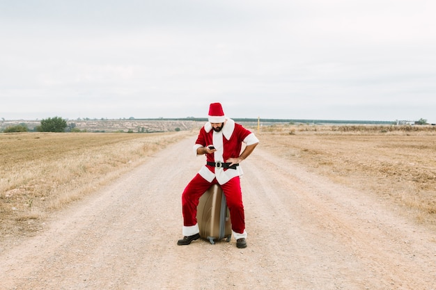 Weihnachtsmann sitzt auf einem Koffer mit einem Handy auf dem Land.