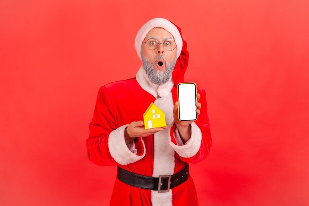 Weihnachtsmann steht mit Papierhaus und Handy mit leerem Bildschirm, Umzugsdienste