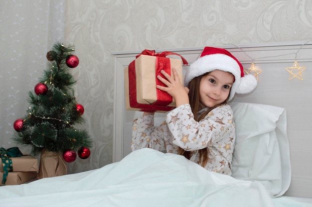 Weihnachtsmorgen, ein kleines Mädchen im Schlafanzug im Bett freut sich mit Geschenken auf dem Hintergrund eines Weihnachtsbaums. Ein glückliches, lächelndes Kind öffnet zu Hause ein Neujahrsgeschenk. Das Konzept der Feiertage, Weihnachten