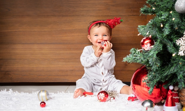 Weihnachtsporträt des Babys auf der Holzwand nahe dem Weihnachtsbaum. Kind mit Weihnachtsverzierung