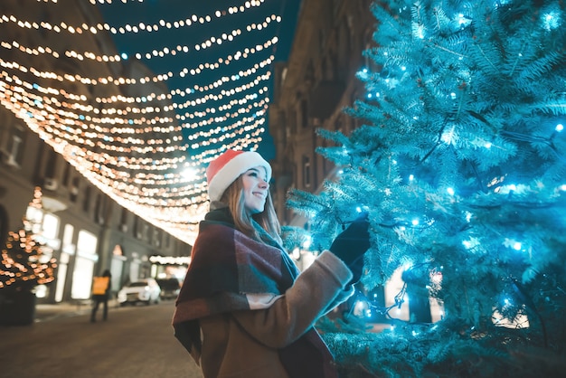 Weihnachtsporträt eines lächelnden Mädchens in warmen Kleidern am Abend auf einer geschmückten Straße in den Lichtern nahe dem Weihnachtsbaum