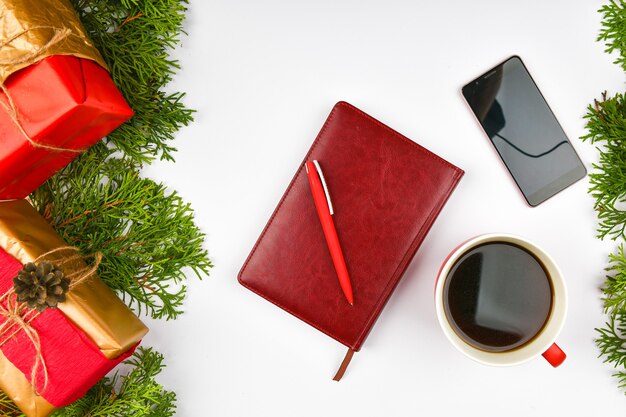 Weihnachtsraum von Notizblock, Telefon, Kaffeetasse. Keramische rote Tasse mit schwarzem Kaffee. Neujahrslayout. Sicht von oben. Eine weibliche Hand schreibt in ein Notizbuch mit Adern.