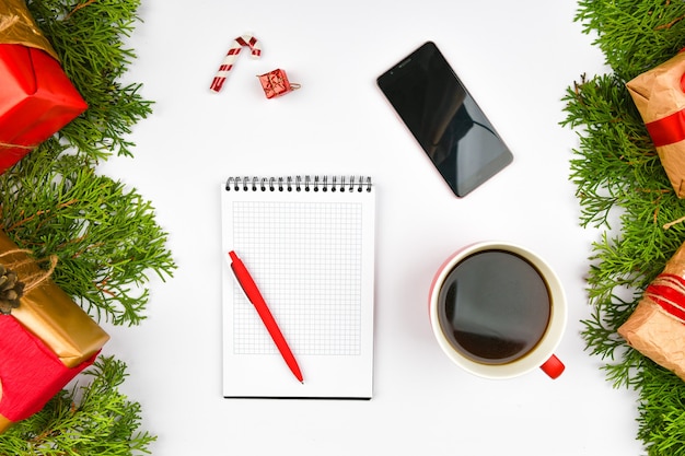 Weihnachtsraum von Notizblock, Telefon, Kaffeetasse. Keramische rote Tasse mit schwarzem Kaffee. Neujahrslayout. Sicht von oben. Eine weibliche Hand schreibt in ein Notizbuch mit Adern.
