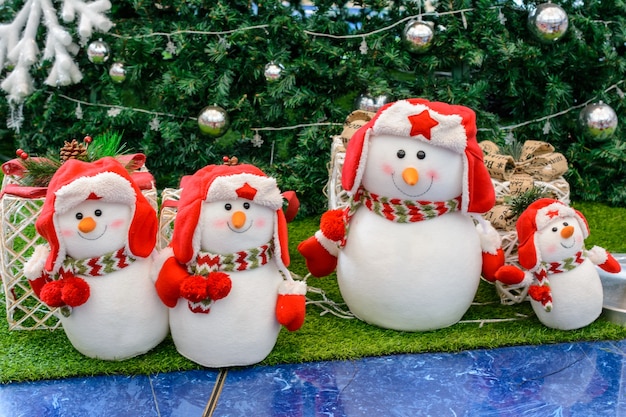 Weihnachtsschneemänner sitzen unter einem Weihnachtsbaum mit Geschenken in roten Kappen Ohrenklappen mit einem Stern, Handschuhen und bunten Schals. Weihnachtskonzept.