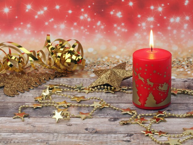 Weihnachtszusammensetzung der brennenden roten Weihnachtskerze, des goldenen Sternentannenbaums und der Girlande auf einer hölzernen Oberfläche und rotem Hintergrund