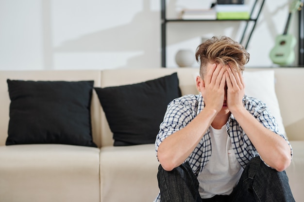 Weinender frustrierter Teenager, der auf dem Boden im Wohnzimmer sitzt und Gesicht mit Händen bedeckt