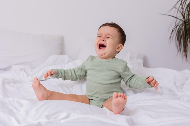 Weinendes Baby in einem grünen Baumwollbody und nackten Füßen sitzt auf einem ausgebreiteten Bett