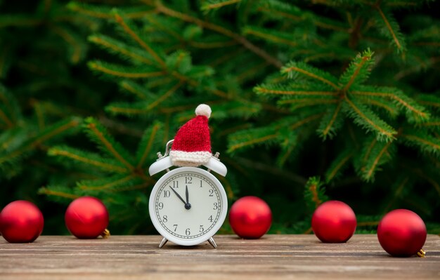 Weinlesewecker mit Weihnachtsmütze und Kugeln auf Holztisch mit Fichtenzweigen auf Hintergrund