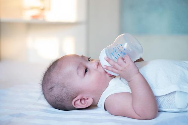 Weiß gekleidetes asiatisches Baby liegt auf dem Bett und trinkt Milch aus einer Flasche