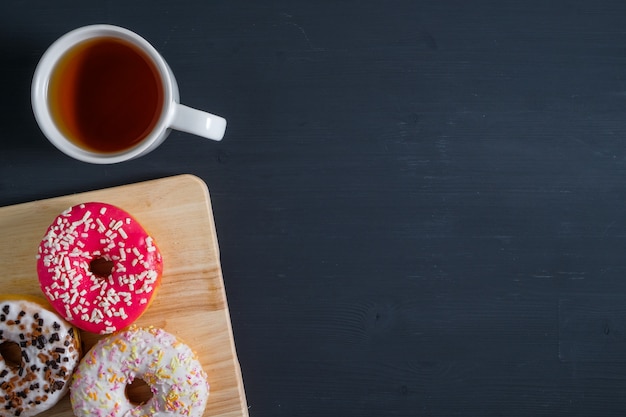 Weiß, rosa und braun glasierte Donuts mit einer Tasse Tee auf der linken Seite auf schwarzem Holz