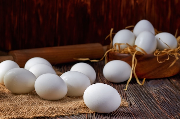 Weiße Eier auf Sackleinen und Holztisch. Im Hintergrund sind Eier in einer Holzschale und ein Nudelholz. Zurückhaltend.