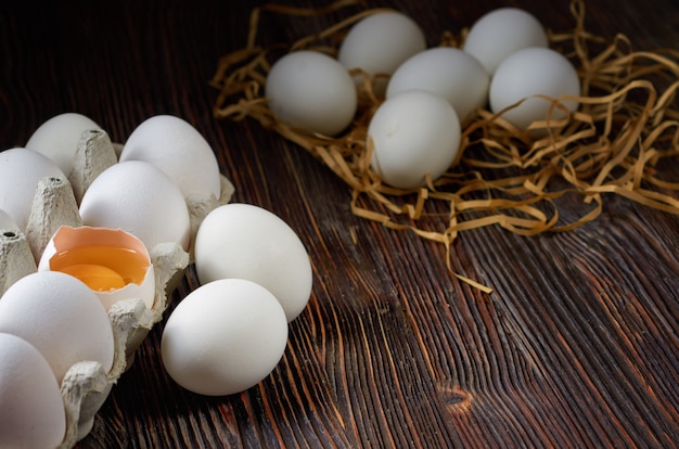 Weiße Eier in einer Packung auf Papierstroh und Holztisch. Gebrochenes Ei mit Eigelb auf der Vorderseite. Zurückhaltend.