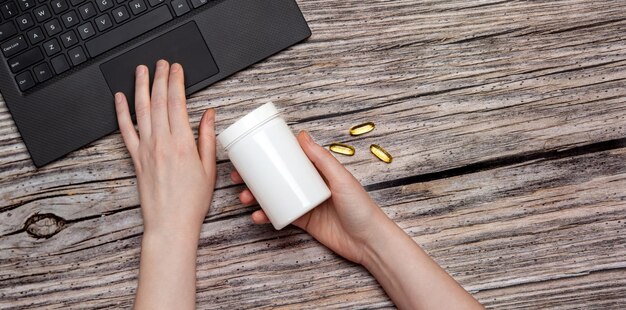 Weiße Flasche mit Omega-Vitaminpillen in der Hand und Laptop auf Holzhintergrund